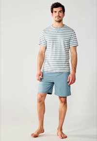 Mann steht barfuß und trägt ein hellblau gestreiftes T-Shirt sowie einfarbige hellblaue Shorts vor einem schlichten Hintergrund.