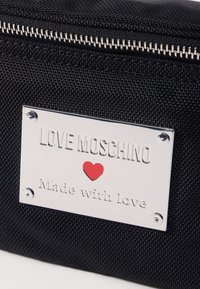 Schwarze Nylon-Tasche mit einer silbernen Logo-Plakette mit erhabenem Text. Ein kleines rotes Herz ist in das Design integriert und sorgt für einen Kontrast.