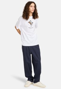 White cotton t-shirt with a central graphic, paired with loose navy trousers and cream sneakers. Straight-cut design and short sleeves.