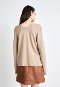 Beige Pullover mit V-Ausschnitt und überschnittenen Schultern, aus weichem Stoff, kombiniert mit einem braunen Lederrock mit geradem Saum.