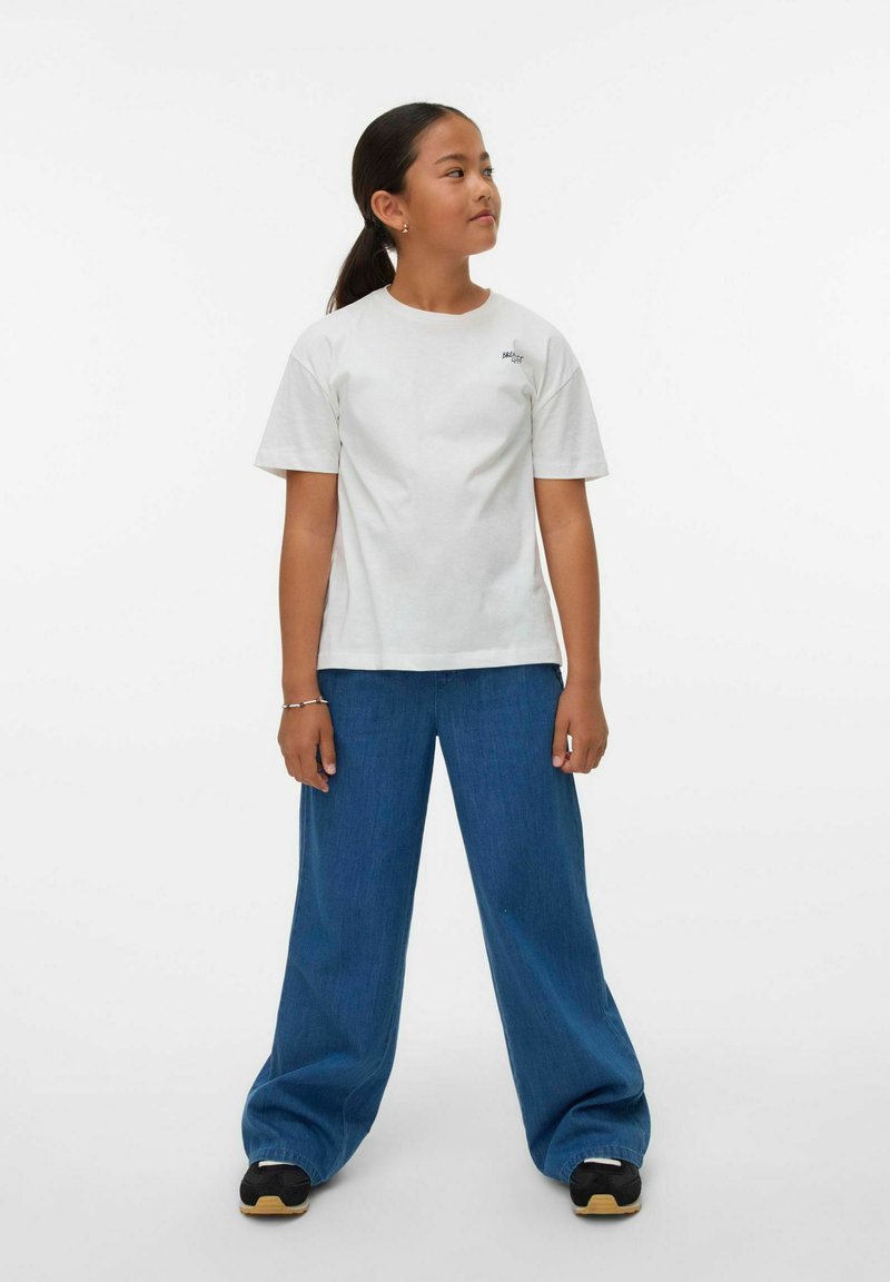 Camiseta de algodón blanca de mangas cortas, combinada con pantalones de mezclilla azul holgados. El conjunto se completa con zapatillas negras con suela beige.