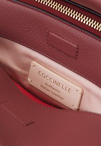 Piros bőrbelső egy táskában, amelyen egy bézs címke olvasható "COCCINELLE Authentic Italian Leather" arany betűkkel. Strukturált felület.