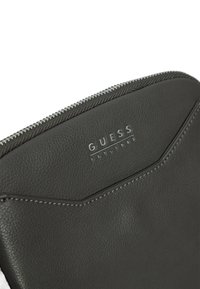 Clutch de imitación de cuero negro con textura, cierre de cremallera plateado y logo de GUESS. Presenta un diseño geométrico con costuras en contraste.