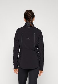 Black jacket with a high collar, two zippered pockets on the back, and tonal stitching. Features a circular logo on the upper back.