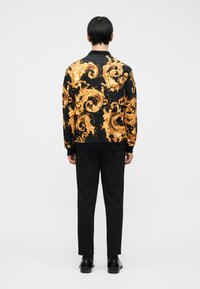 Άτ ομο φαίνεται από πίσω φορώντας μαύρο μπουφάν με περίτεχνα χρυσά κυματιστά σχέδια, μαύρο παντελόνι και μαύρα παπούτσια φόρμας, πάνω σε απλό φόντο.