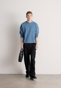 Καθημερινό μπλε πουλόβερ, άνετη εφαρμογή, μακριά μανίκια, σε συνδυασμό με μαύρο παντελόνι. Το μοντέλο κρατά μια μικρή μαύρη τσάντα, στέκεται σε λεία επιφάνεια.