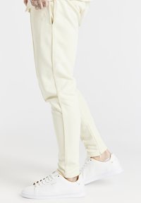 Fittowane spodnie dresowe w kolorze kremowym z bocznymi kieszeniami, w połączeniu z białymi sneakersami z złotymi akcentami i logo. Gładka faktura i smukły krój.
