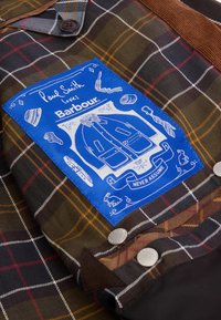 Sötétbarna, zöld és piros árnyalatú kockás kabát, elülső részén kék címkével, fehér illusztrációkkal és ezüst gombokkal.