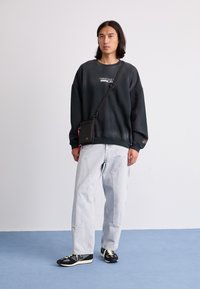 Felpa nera oversize con logo, jeans baggy blu chiaro e sneakers nere con dettagli bianchi. Il modello è in piedi contro uno sfondo semplice.