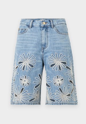 Nő áll, kezét a zsebében tartja, fehér ujjatlan pólót és térd alá érő farmer rövidnadrágot visel, melyeken nagy hímzett virágminták vannak.