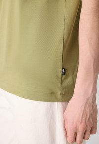 Olíva zöld T-shirt sima textúrával, oldalsó hasítékkal és kis fekete 'BOSS' logó címkével az alsó szegélyen, fehér nadrággal párosítva.