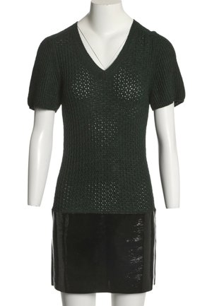Haut en maille noire à manches courtes avec col en V, associé à une mini-jupe noire brillante sur un mannequin blanc sur fond blanc.