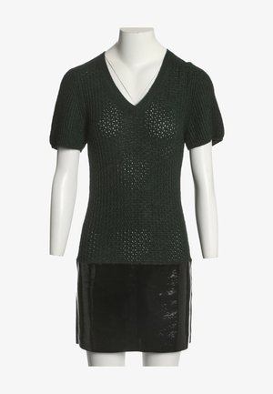 Haut en maille noire à manches courtes avec col en V, associé à une mini-jupe noire brillante sur un mannequin blanc sur fond blanc.