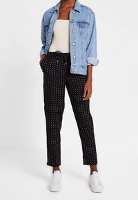 Veste en denim bleu clair, portée sur un haut blanc ajusté. Pantalon noir à pois blancs avec cordon de serrage, associé à des baskets blanches.