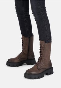 Brown leather lace-up boots with a chunky black sole, side zipper, and textured surface. Features metal eyelets and reinforced stitching.
