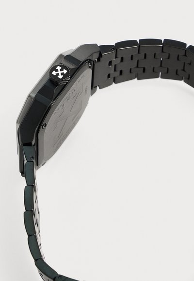 Bracelet de montre en métal noir avec un boîtier carré et une couronne arborant un logo blanc à quatre flèches sur fond clair.