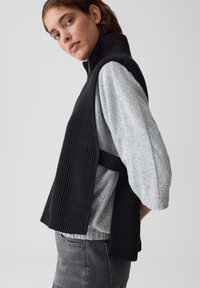 Frau, die ein schwarzes geripptes ärmelloses Top über einem hellgrauen Pullover und dunkle graue Jeans trägt, und vor einem schlichten hellen Hintergrund posiert.