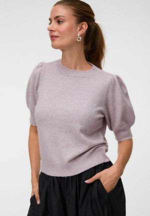 Femme aux cheveux bruns en queue de cheval, portant des boucles d'oreilles argentées, un pull court à manches bouffantes violet clair et une jupe noire, les mains dans les poches.