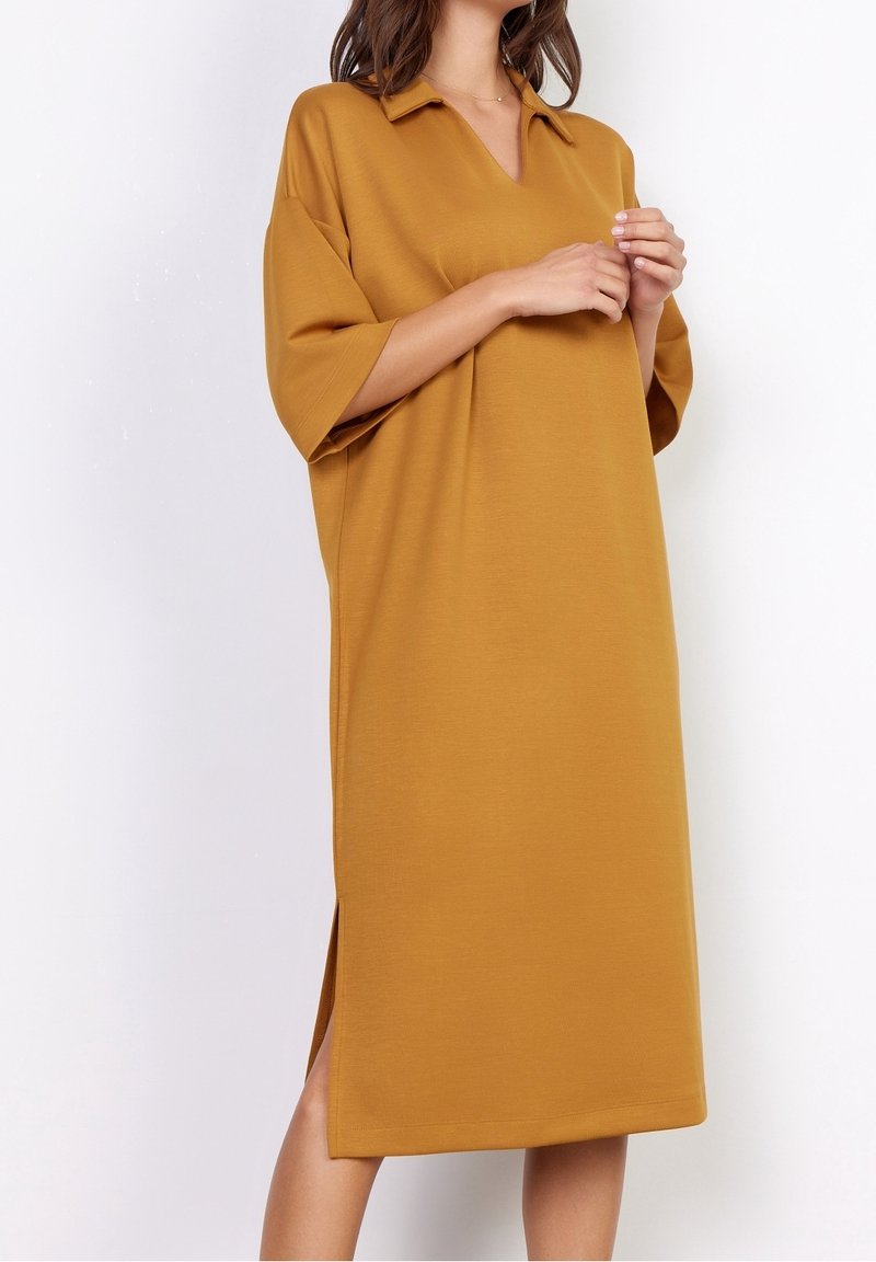 Femme portant une robe ample jaune moutarde longueur genou avec manches courtes, col et fentes latérales, debout devant un fond blanc.