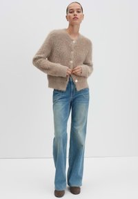 Cardigan beige moelleux avec cinq boutons, associé à un jean évasé bleu clair et des bottines brunes. Texture lisse et coupe décontractée.