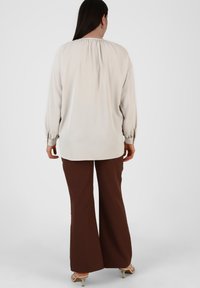 Blusa lunga color beige chiaro con scollatura arricciata, abbinata a pantaloni larghi marroni e sandali con tacco beige. Texture liscia.