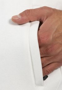 Un tejido blanco con una textura suave presenta una mano posicionada en un bolsillo lateral. Un pequeño detalle negro es visible cerca de la apertura del bolsillo.