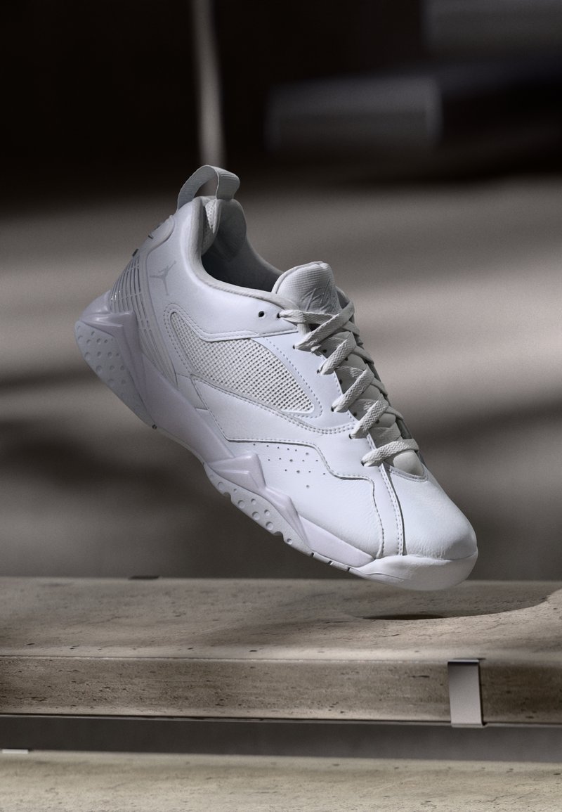 Weißer Sportschuh mit Netzpaneelen und Schnürsenkeln, über einer Betonbank in weichem Licht und Schatten schwebend.