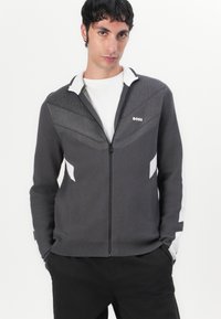 Graue Zip-Jacke mit weißen Akzenten, die ein strukturiertes dreieckiges Muster auf der Brust und eine LOGO-Details auf der linken Seite aufweist.