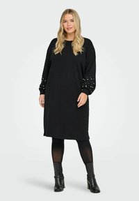 Robe noire jusqu'aux genoux en tissu doux avec des manches longues. Les poignets des manches sont ornés de clous blancs décoratifs. Portée avec des bottes noires et des collants.