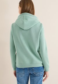 Sweat à capuche vert menthe avec une capuche imposante, des poignets et un ourlet côtelés. La vue de dos montre une texture de tissu lisse et une coupe carrée, complétée par un jean bleu.