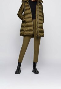 Olivově zelený prošívaný kabát s kapucí, vybavený zipovým zapínáním, žebrovanými manžetami a bočními kapsami. Kombinován se sladěnými olivovými kalhotami a černými botami.