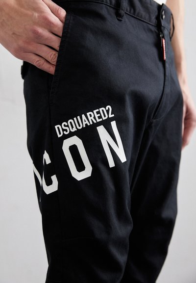 Black pants made of fabric with a smooth texture featuring white "DSQUARED2 ICON" lettering on the side and a hand in the pocket.