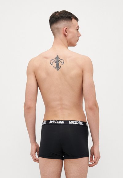 Black boxer briefs with a white Moschino logo waistband; model has a tattoo on his back, showcasing smooth skin and a fitted design.