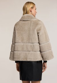 Žena oblečená v béžové kráčené umělé kožešině s širokými rukávy a černé kožené sukni, zobrazená zezadu na neutrálním pozadí.