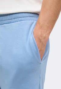 Gros plan sur la main d'une personne reposant dans la poche d'un pantalon de jogging bleu clair avec une chemise blanche légèrement rentrée au-dessus de la ceinture.