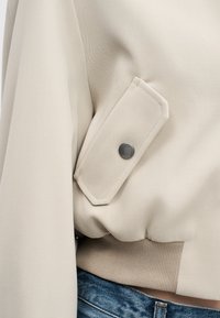 Veste beige avec une poche à rabat à boutons-pression et un ourlet côtelé, portée sur un jean en denim bleu.