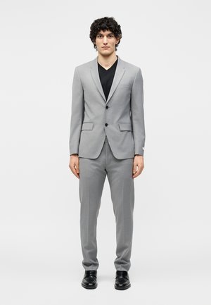 Νέος άνδρας με σγουρά μαλλιά που φοράει ανοιχτό γκρι κοστούμι, μαύρο πουκάμισο με V-λαιμόκοψη και μαύρα επίσημα παπούτσια, στέκεται μπροστά σε απλό λευκό φόντο.