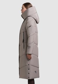 Lang, puffy beige frakke med høj krave og hætte. Har lynlåse, trykknapper og et logo-patch; det kviltede design tilføjer tekstur.