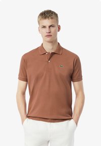 Brun bomullspiké med korta ärmar, krage, tvåknapplåsning och en grön logga på vänster bröst. Avslappnad passform.