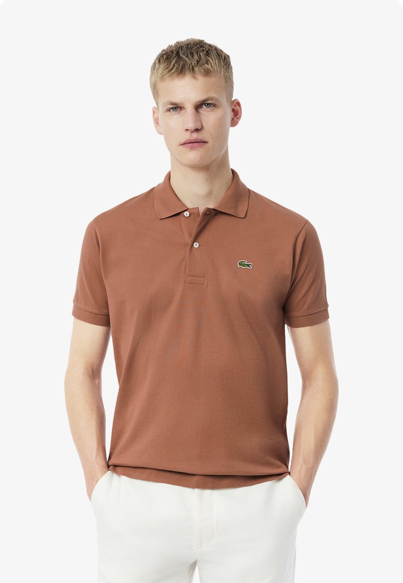 Brun bomullspiké med korta ärmar, krage, tvåknapplåsning och en grön logga på vänster bröst. Avslappnad passform.