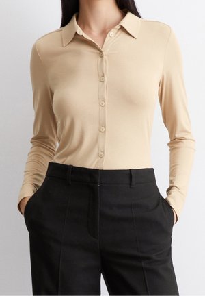 Femme portant une chemise beige à manches longues boutonnée, rentrée dans un pantalon noir taille haute ajusté, mains dans les poches.
