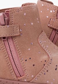 Botines de ante color tan con cremalleras rosas, que presentan motas brillantes en contraste. Punta redondeada y detalles cosidos en los laterales.