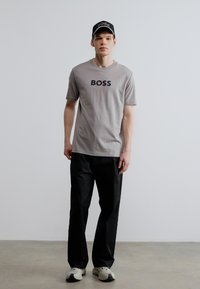 Γκρι μπλουζάκι με την επιγραφή "BOSS" σε μαύρο, συνδυασμένο με μαύρο παντελόνι και λευκά αθλητικά παπούτσια. Το μοντέλο φορά επίσης μαύρο καπέλο.