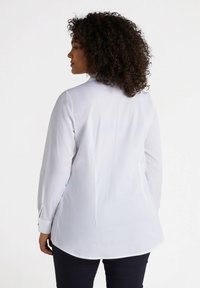 Camicia bianca a maniche lunghe con colletto classico, orlo curvo e polsini bottonati, realizzata in tessuto liscio. Indossata con pantaloni blu navy.