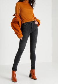 Jeans ajustados negros combinados con un suéter de cuello alto naranja texturizado y una chaqueta naranja acolchada. El conjunto se complementa con botas de tacón naranja brillantes.