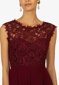 Das burgunderfarbene Kleid verfügt über ein florales Spitzenoberteil mit einem transparenten Overlay, einem gewellten Ausschnitt und einem glatten Rockteil. Es ist mit großen Ohrringen akzentuiert.