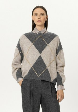 Woman with straight dark hair wearing a grey and beige argyle sweater over a light gray shirt and dark gray pleated pants with hands in pockets.