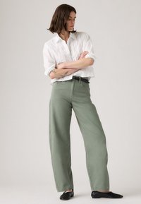 Femme aux cheveux bruns portant une blouse blanche et un pantalon large vert olive, les bras croisés, se tenant devant un fond clair.