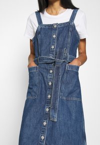 Sukienka ogrodniczka denimowa z regulowanymi ramiączkami, przednimi guzikami, bocznymi kieszeniami i materiałowym paskiem. Ciemnoniebieski kolor z delikatną wyblakłą teksturą.