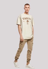 Beige oversized t-shirt with "TAHITI 2023" print, paired with tan cargo pants featuring side pockets, and white sneakers.